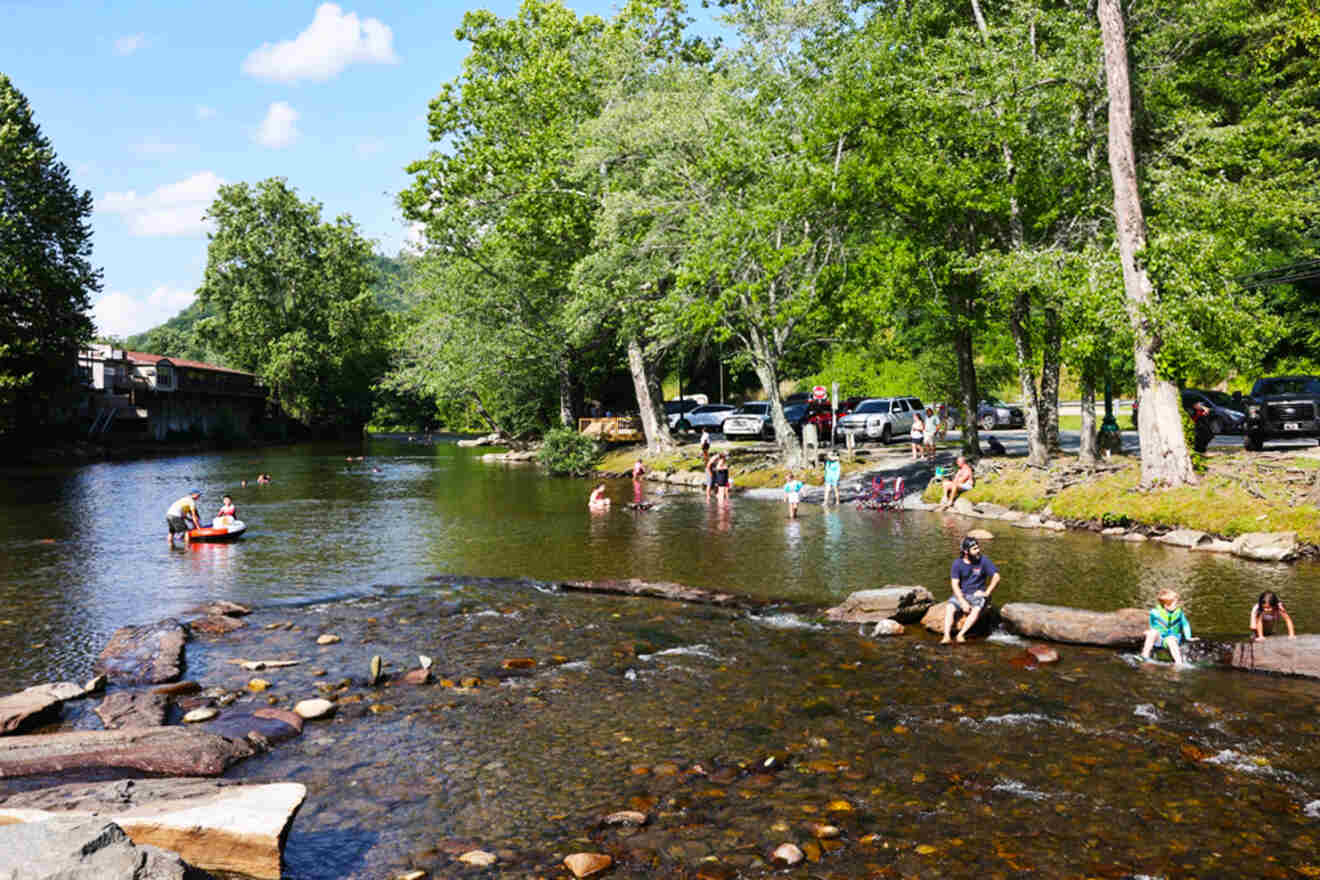 People enjoying a sunny day at a shallow river, with children and adults wading and playing. Trees and parked cars line the riverbank.