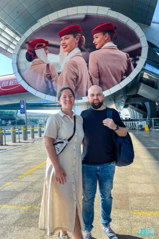 A man and a woman stand smiling outside an airport. Behind them is a large advertisement featuring two flight attendants in uniform.