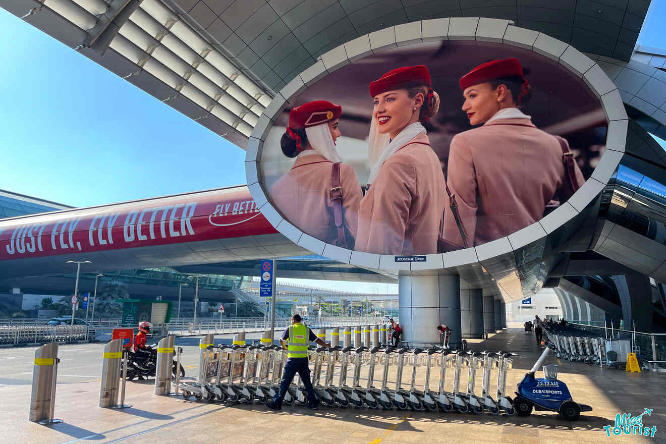 Airport terminal with luggage carts and a large billboard featuring two flight attendants in red uniforms.