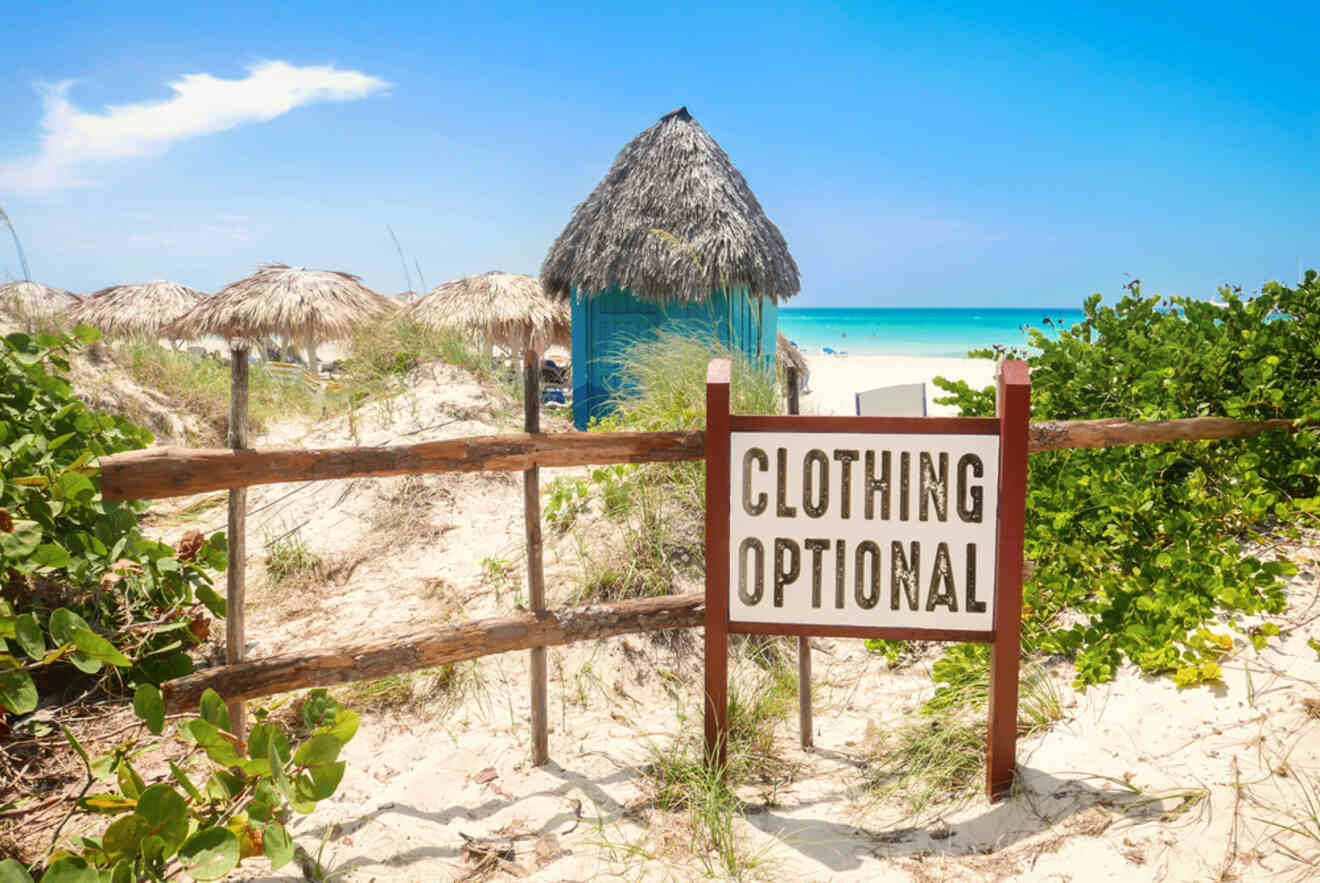 Sign reading "Clothing Optional" in front of a beach scene with sandy dunes, thatched huts, and clear turquoise water under a bright blue sky.