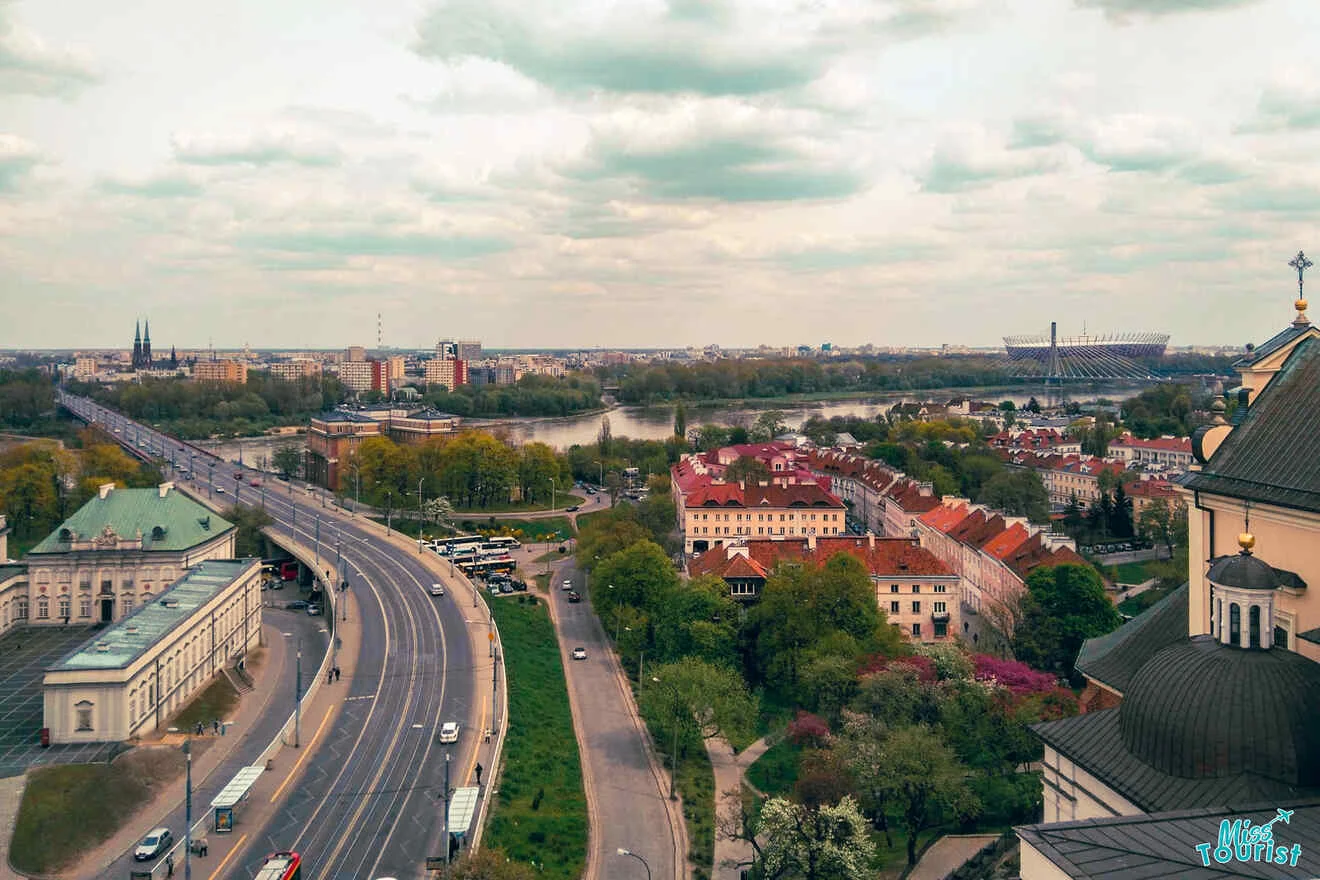 Aerial view of a cityscape with roads, green spaces, and buildings under a cloudy sky.