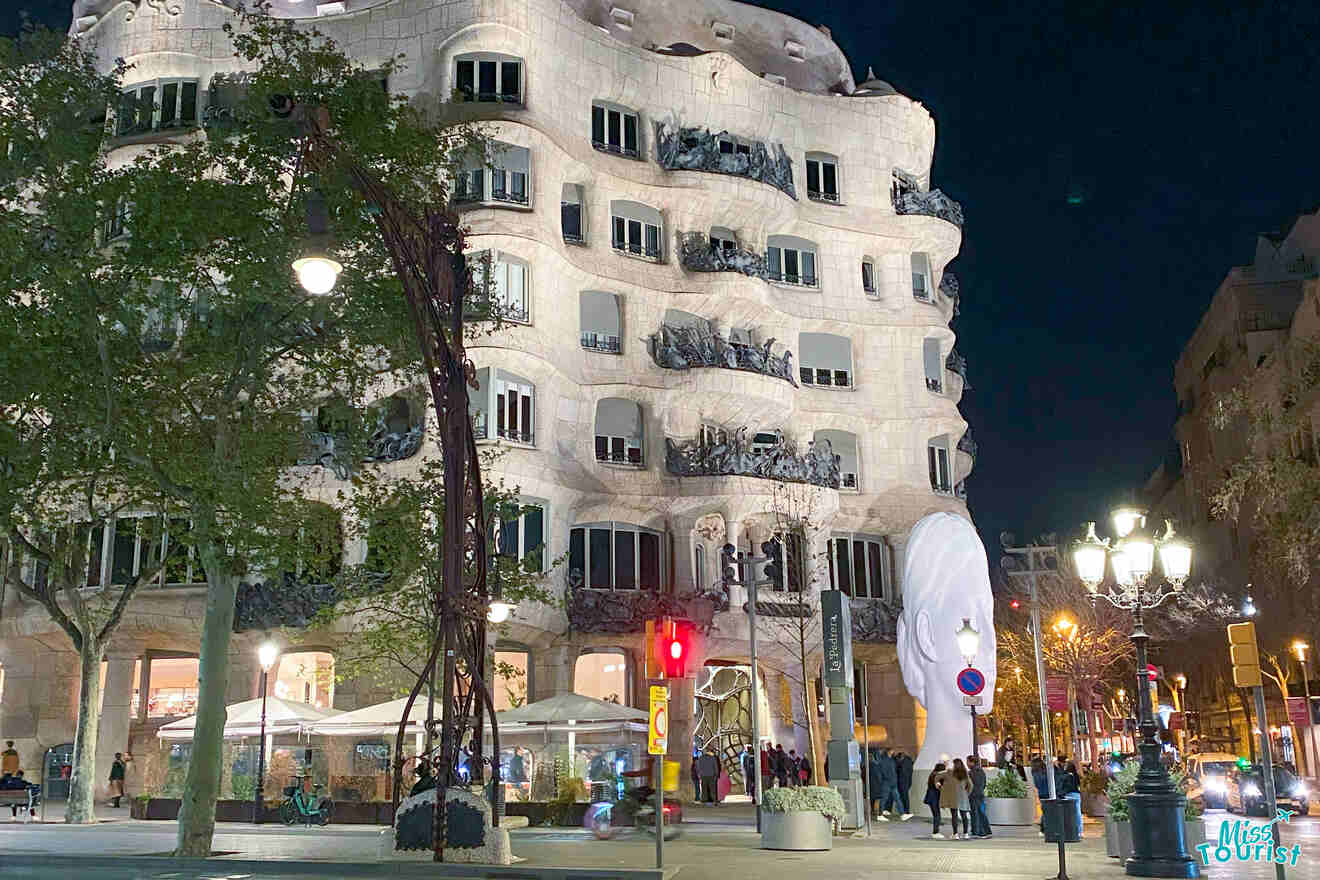 Night view of Casa Milà, a modernist building in Barcelona, with a curving facade, illuminated street lamps, and trees in the foreground. People are gathered near the intersection.