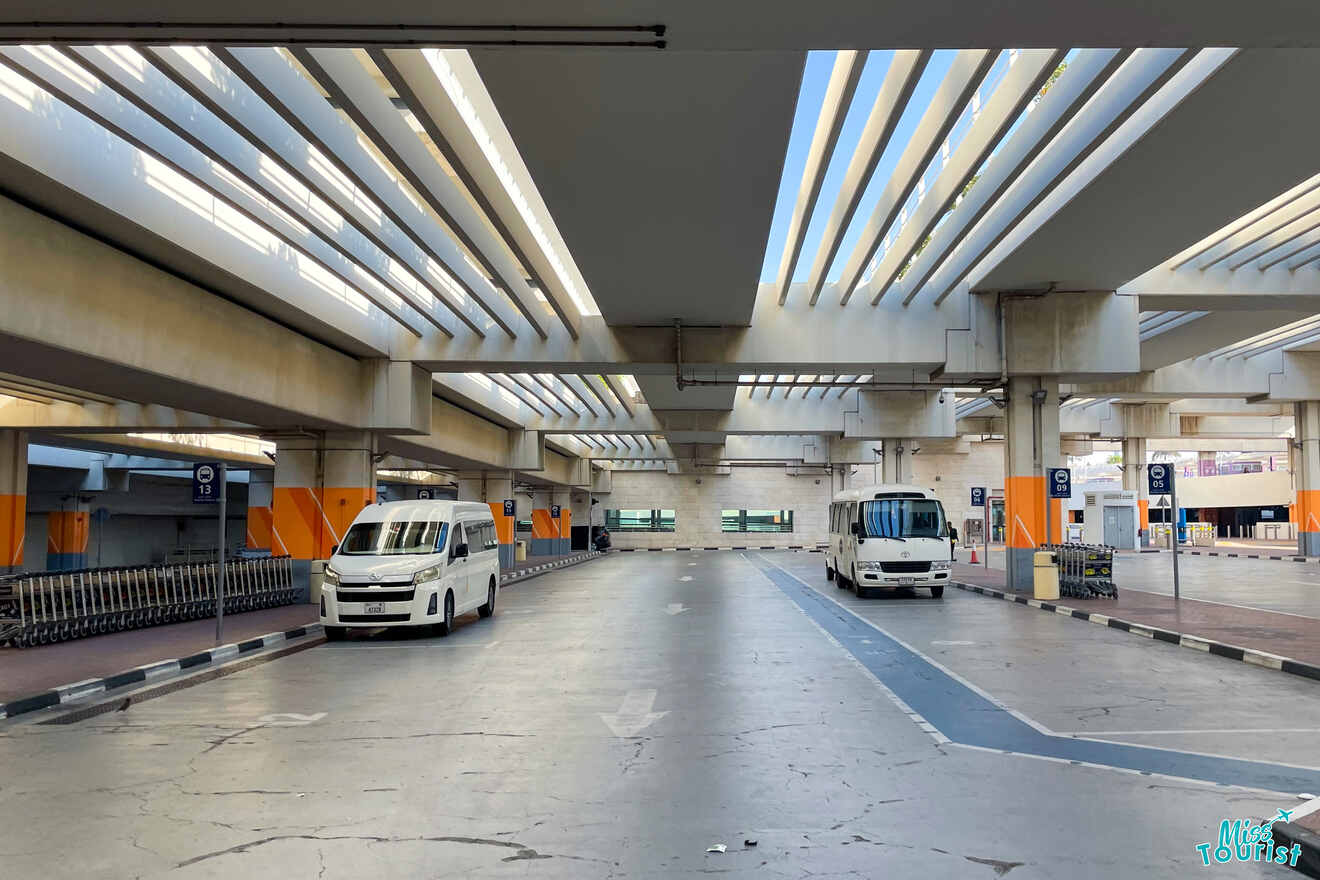 A nearly empty airport shuttle area with two parked buses, a row of luggage carts, and numbered signs hanging from the ceiling.