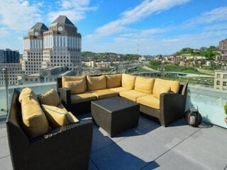 Outdoor rooftop patio with tan cushioned wicker furniture and glass railing, city buildings and greenery visible in the background under a blue sky.