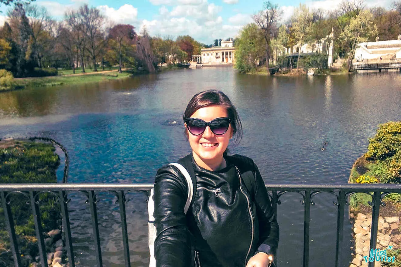 The writer of the post in sunglasses takes a selfie on a bridge over a pond in a park, with trees and a building in the background on a sunny day.