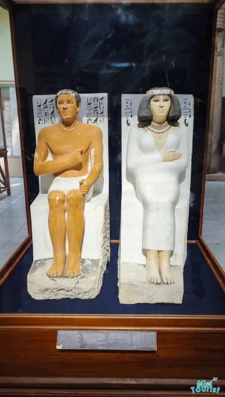 Ancient Egyptian statues of a seated man and woman with hieroglyphs in a display case.