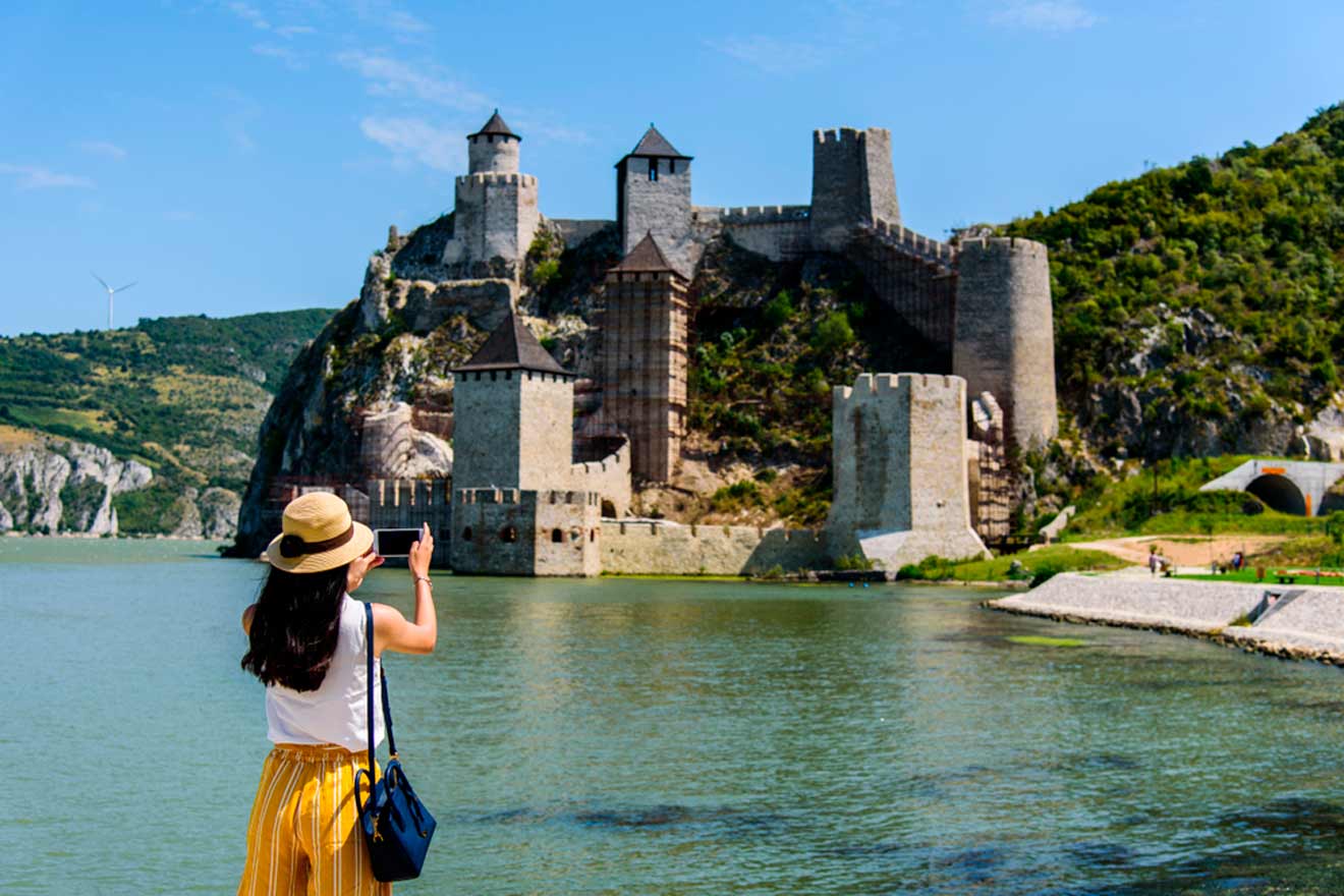 A person in a hat and yellow skirt takes a photo of a medieval fortress by a river under a clear blue sky.