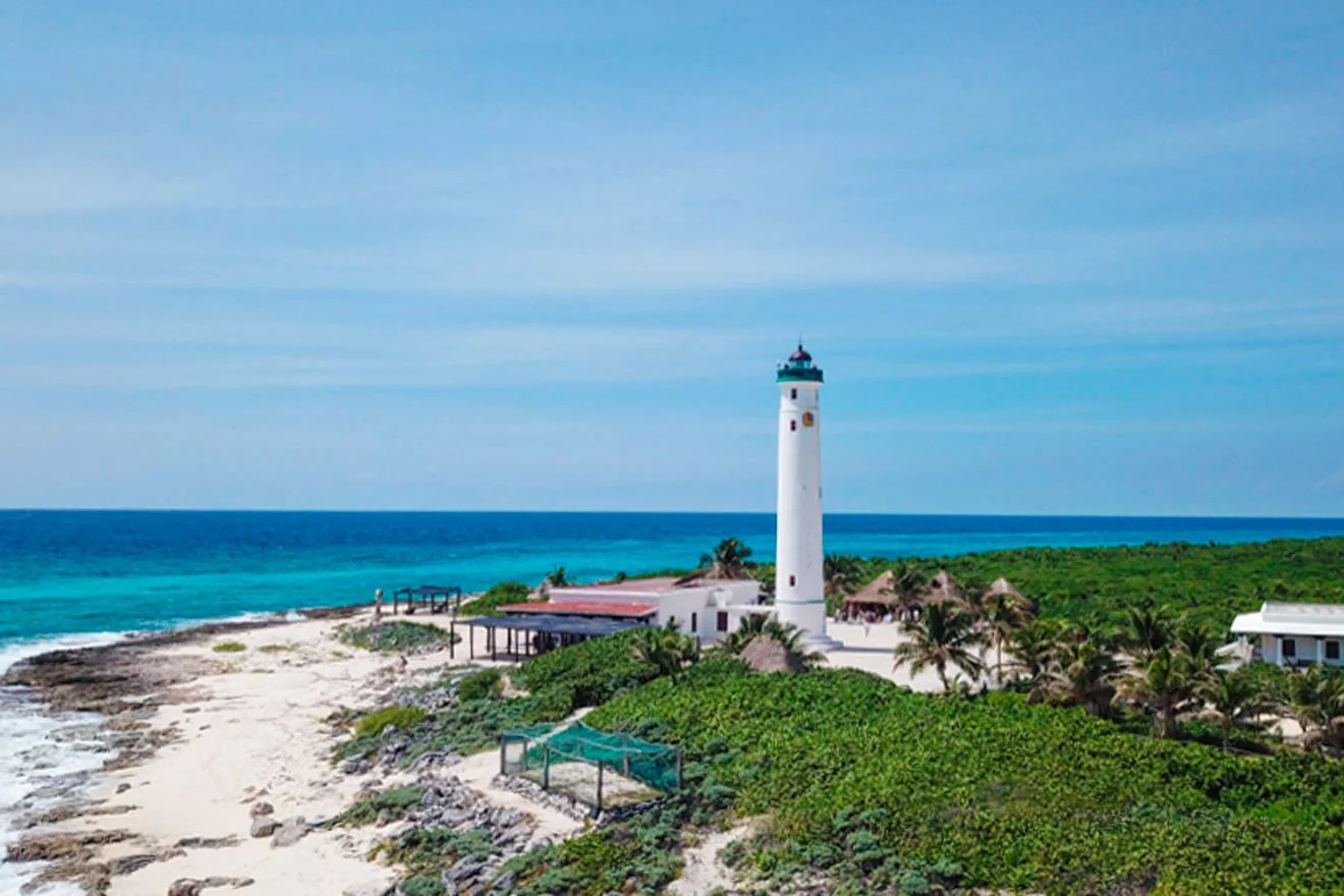 A tall lighthouse stands on a coastal area, surrounded by greenery and a few buildings, with a clear blue sky and ocean in the background.