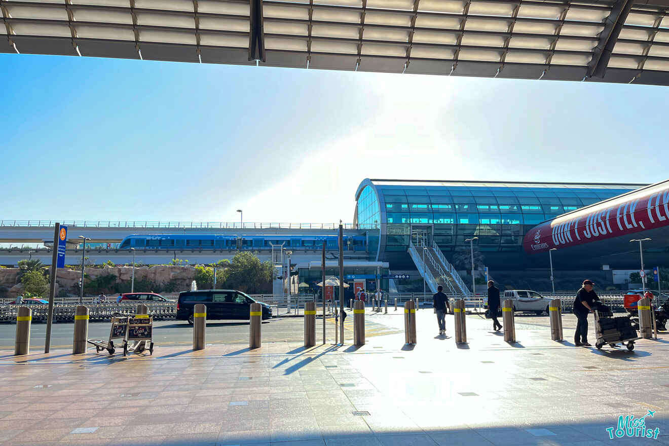 A modern train station with a blue train passing above. People walk under a covered area on a sunny day, with some pushing luggage carts.