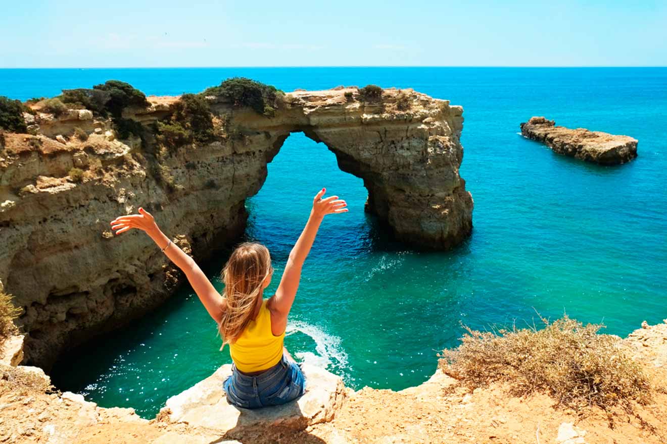 Person sitting on a cliff's edge with arms raised, overlooking a natural rock arch in the ocean under a clear blue sky.