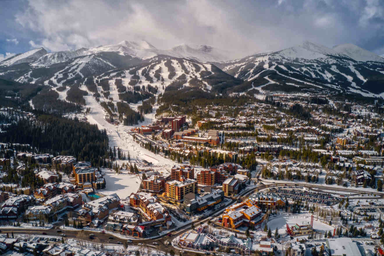 Aerial view of a snowy mountain resort town with ski slopes, buildings, and surrounding forest.