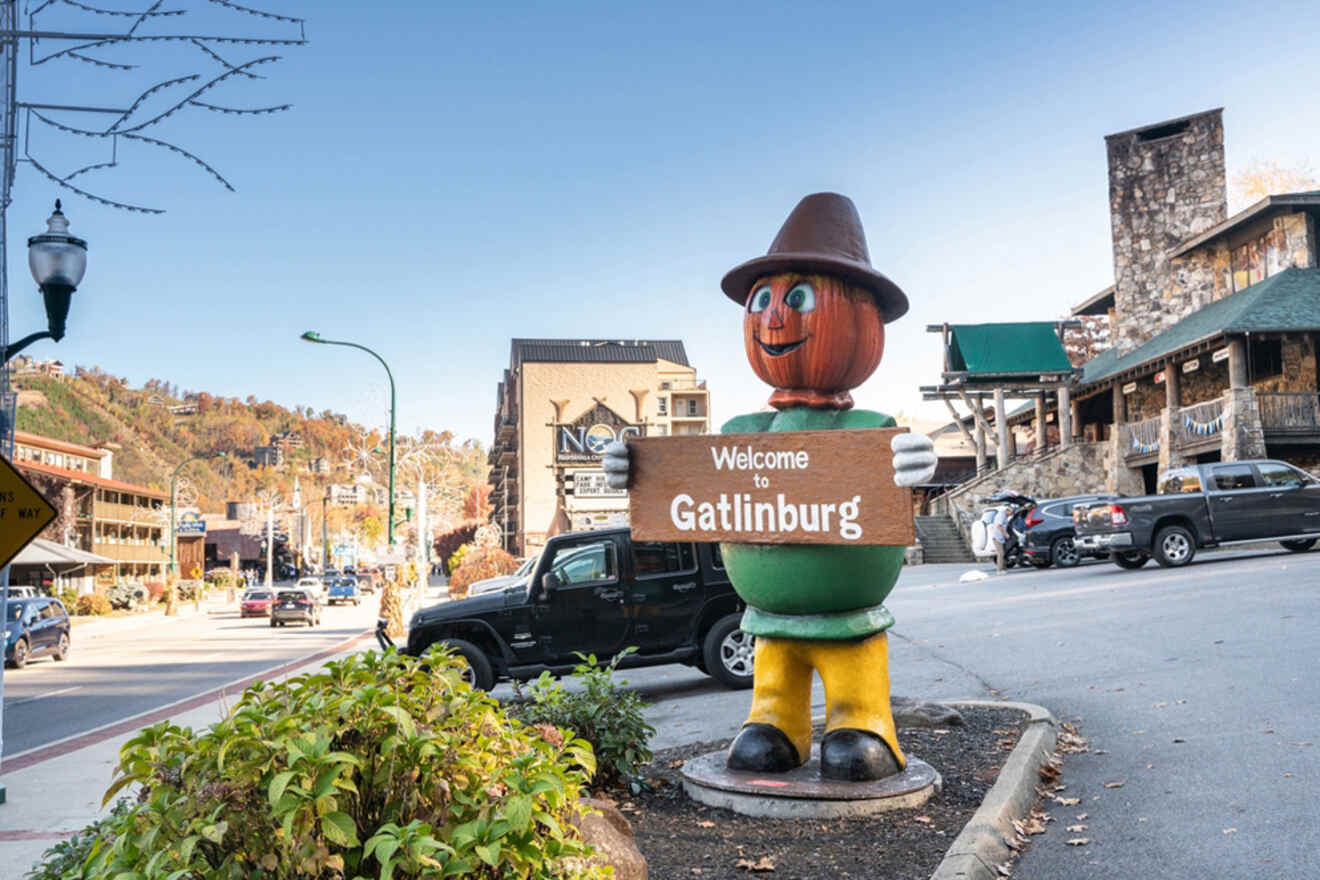 A pumpkin-headed figure holds a "Welcome to Gatlinburg" sign on a busy street with shops and trees in the background.