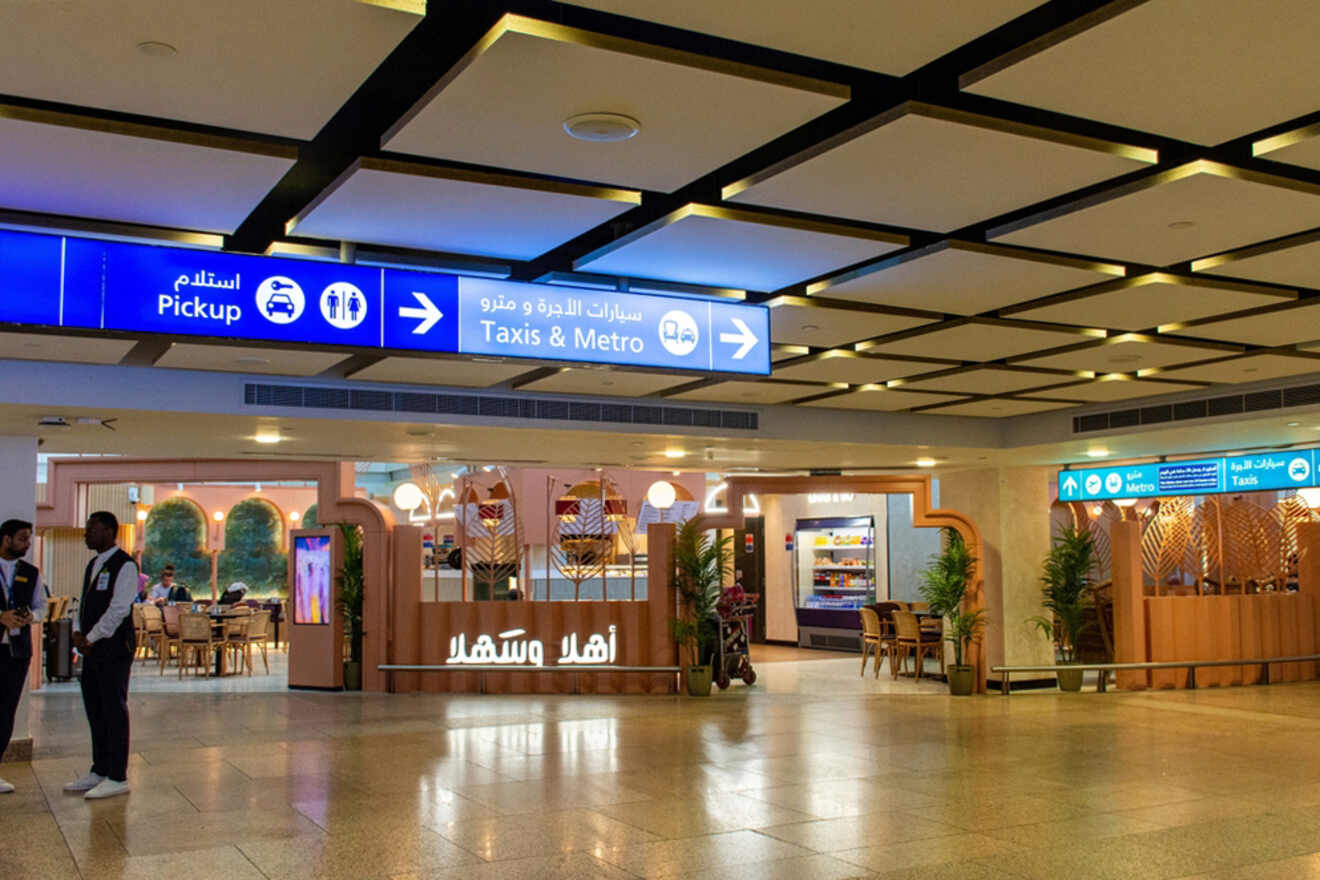 An airport terminal with signs for taxis, metro, and pickups. Shops and people are visible. Ceiling lights are prominent.