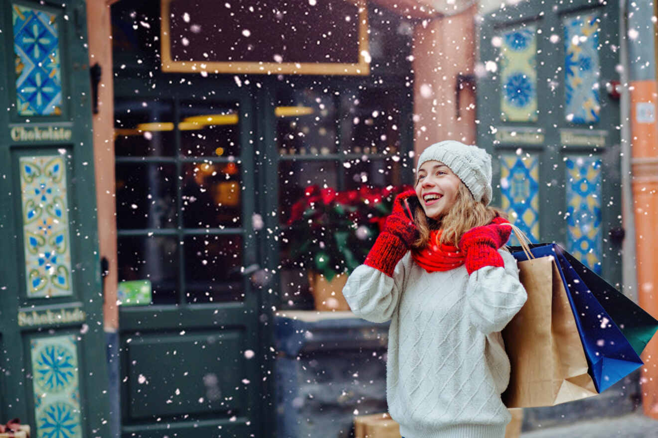 Woman in winter clothing smiles while holding shopping bags during snowfall in a decorated outdoor setting.