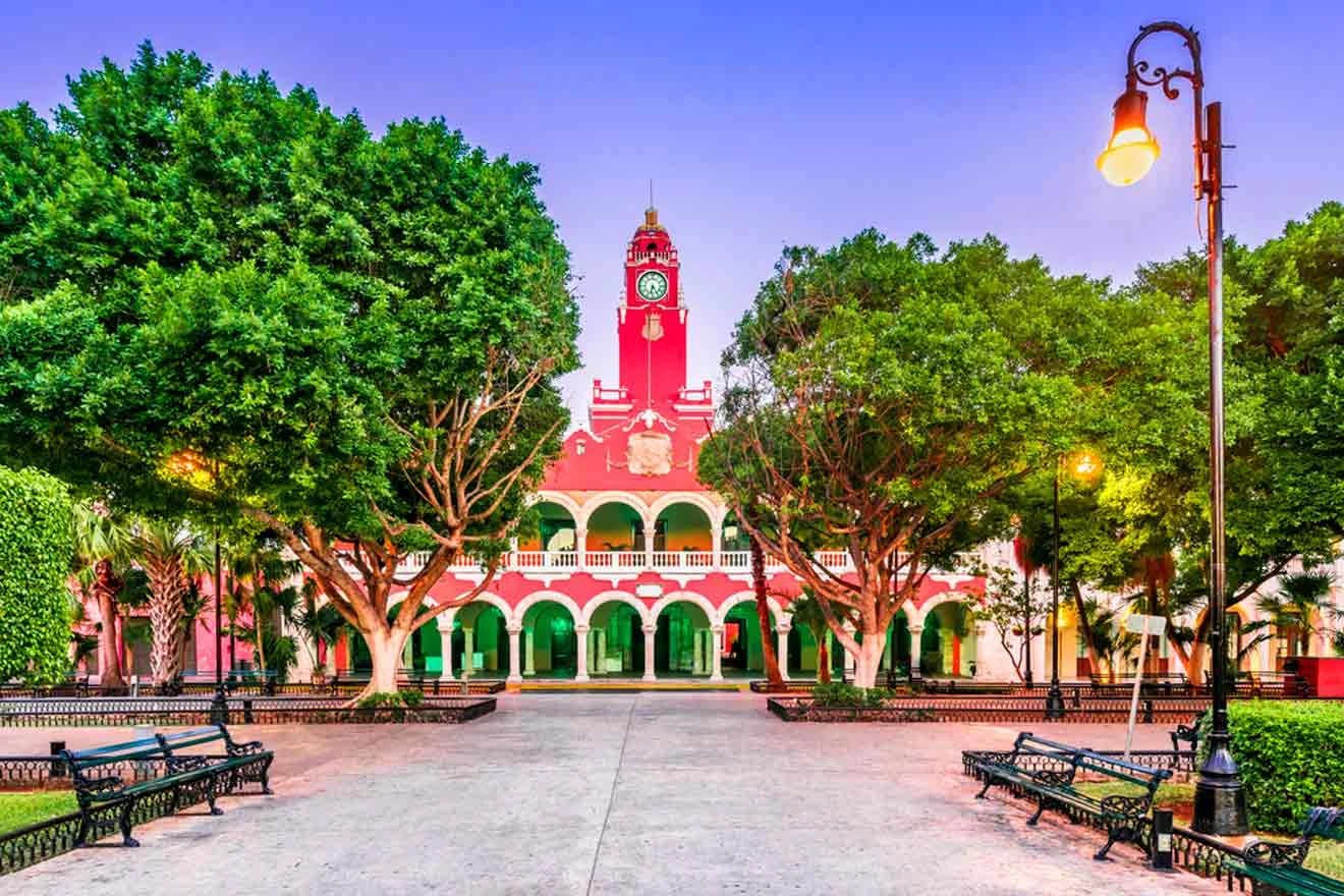 Colonial-style building with arches and a clock tower, surrounded by trees, benches, and streetlights in a park setting.