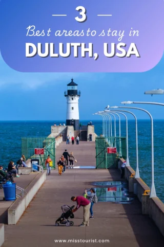 People walking on a pier towards a lighthouse in Duluth, USA, under a clear blue sky.