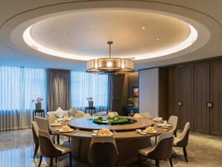 A modern dining room with a large round table, elegant chairs, a central light fixture, and floor-to-ceiling windows with sheer curtains.