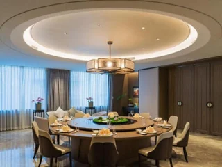 A modern dining room with a large round table, elegant chairs, a central light fixture, and floor-to-ceiling windows with sheer curtains.