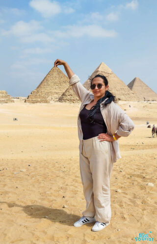 The writer of the post posing in front of the pyramids, wearing sunglasses and light clothing, standing on sandy terrain.