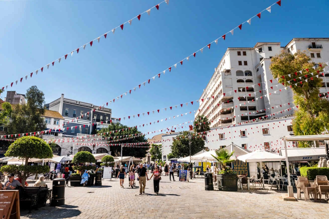 A bustling plaza with people, strung with red and white triangle flags, surrounded by trees, cafes, and multi-story buildings under a clear blue sky.