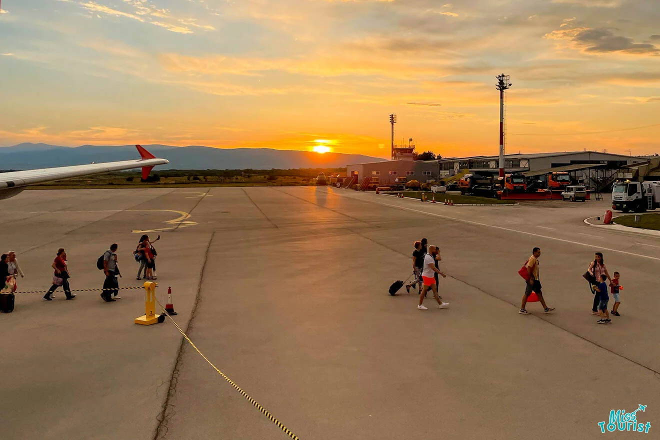 Passengers walking on an airport tarmac at sunset, with a plane and airport buildings in the background.