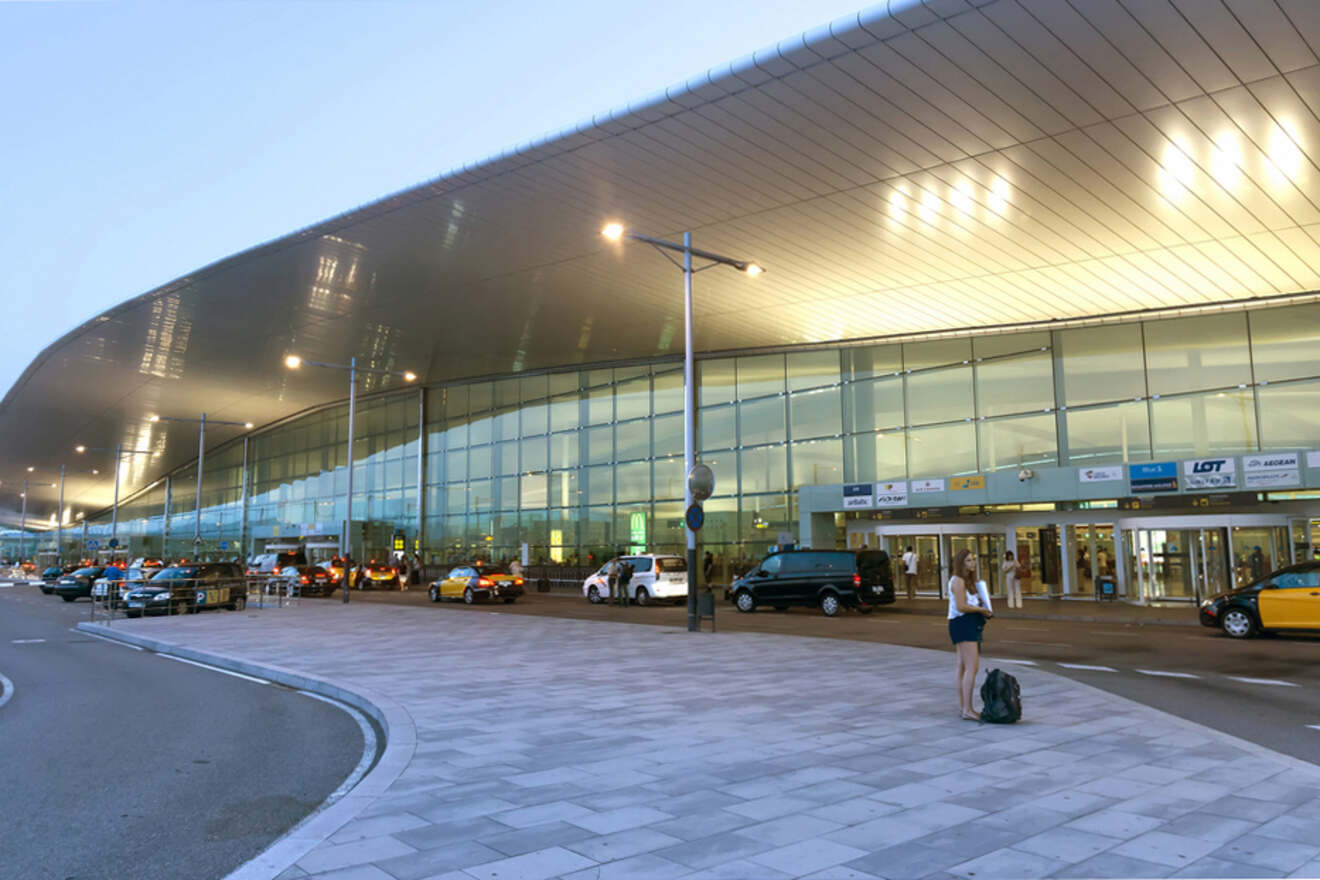 A woman with luggage stands outside a modern airport terminal with taxis and cars parked along the curb.