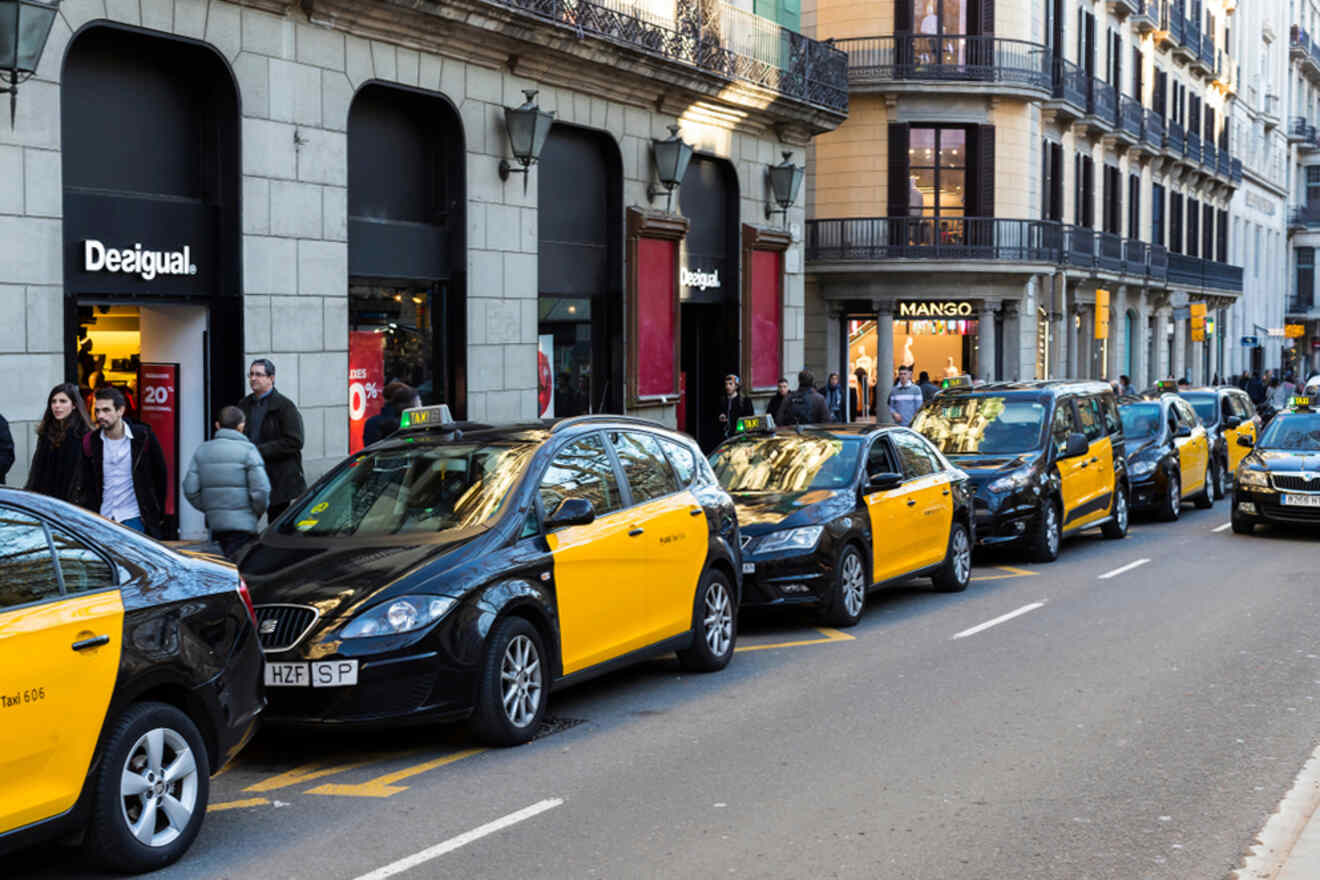 A line of yellow and black taxis parked on a city street near Desigual and Mango stores. Pedestrians walk nearby.