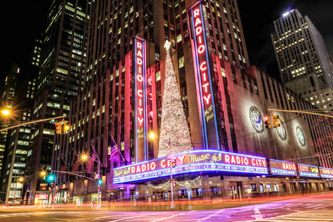 Radio City Music Hall at night, illuminated with bright lights, featuring a large Christmas tree in front. Traffic streaks by, highlighting a city intersection.