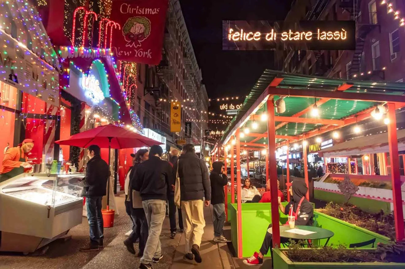 Bustling street scene at night with festive lights, outdoor dining, people walking, and colorful signs in various languages.