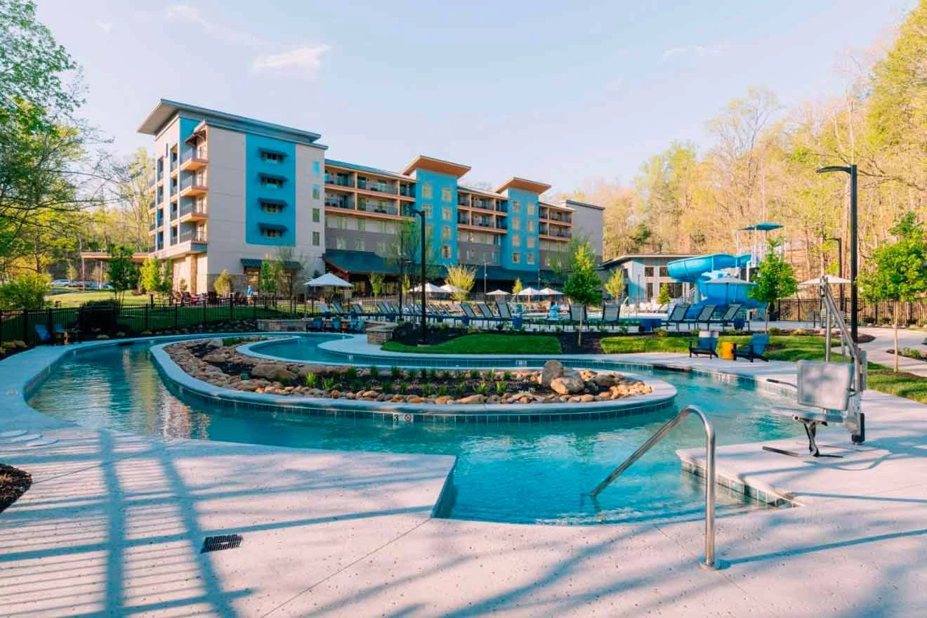 A resort with a lazy river, pool, and waterslide in the foreground. In the background, there is a multi-story building surrounded by trees.