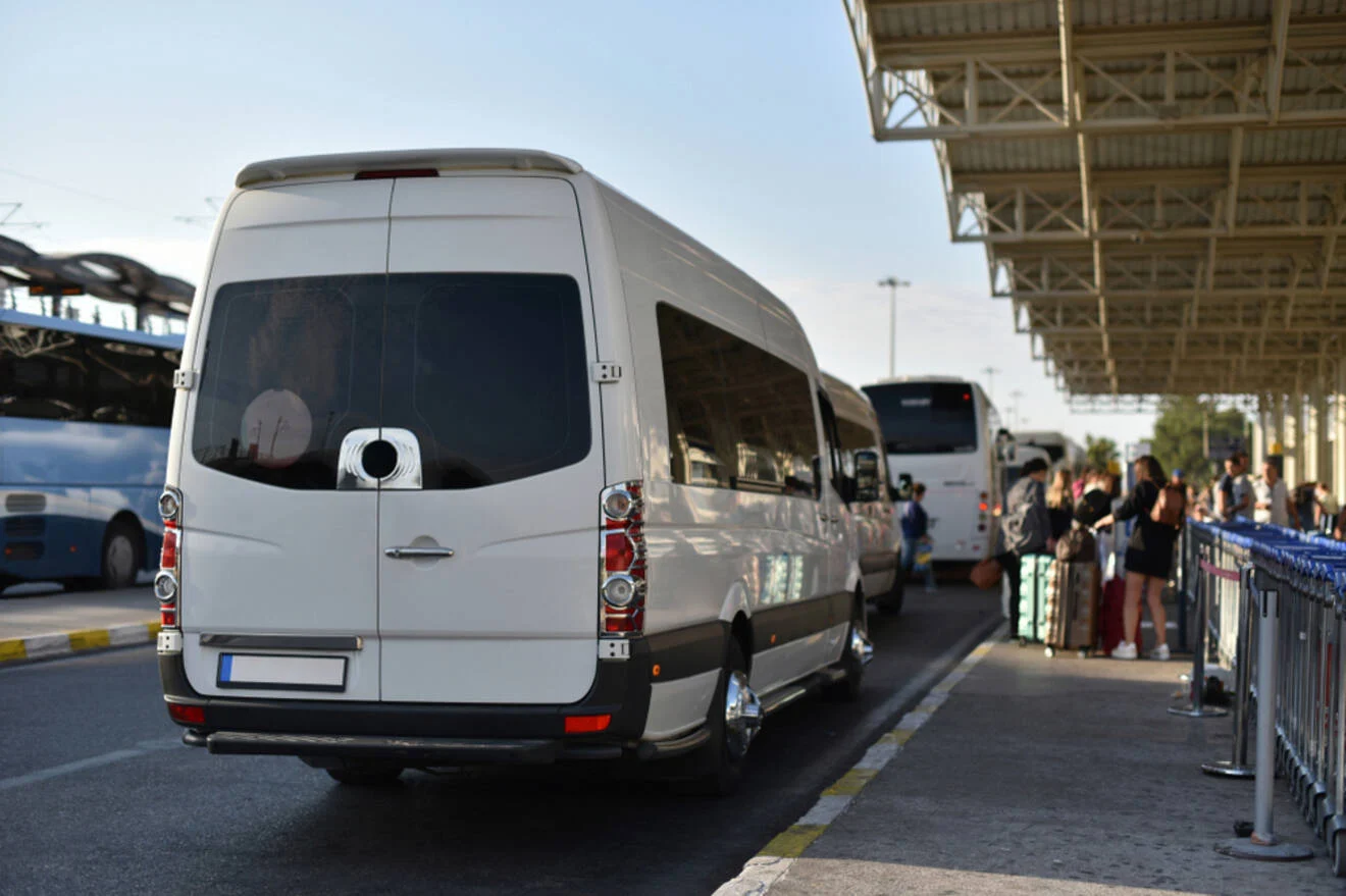 A white passenger van is parked at a bus station next to a line of trolleys. People are visible in the background, some standing near the curb with luggage.