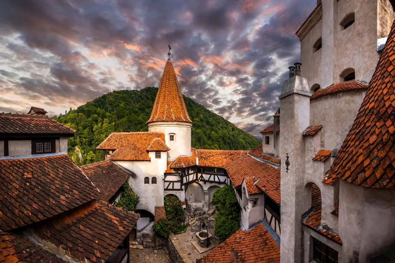 Courtyard of a castle with red-tiled roofs and a central tower against a backdrop of wooded hills and a dramatic sunset sky.