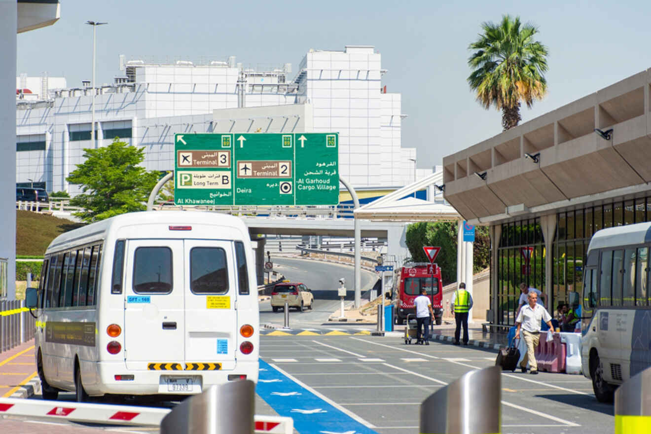 Shuttle buses and people are outside an airport terminal with directional signs visible overhead.