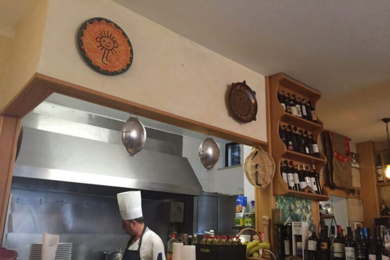 A chef wearing a tall white hat works in a restaurant kitchen; wine bottles and decorative plates are displayed on shelves and walls.