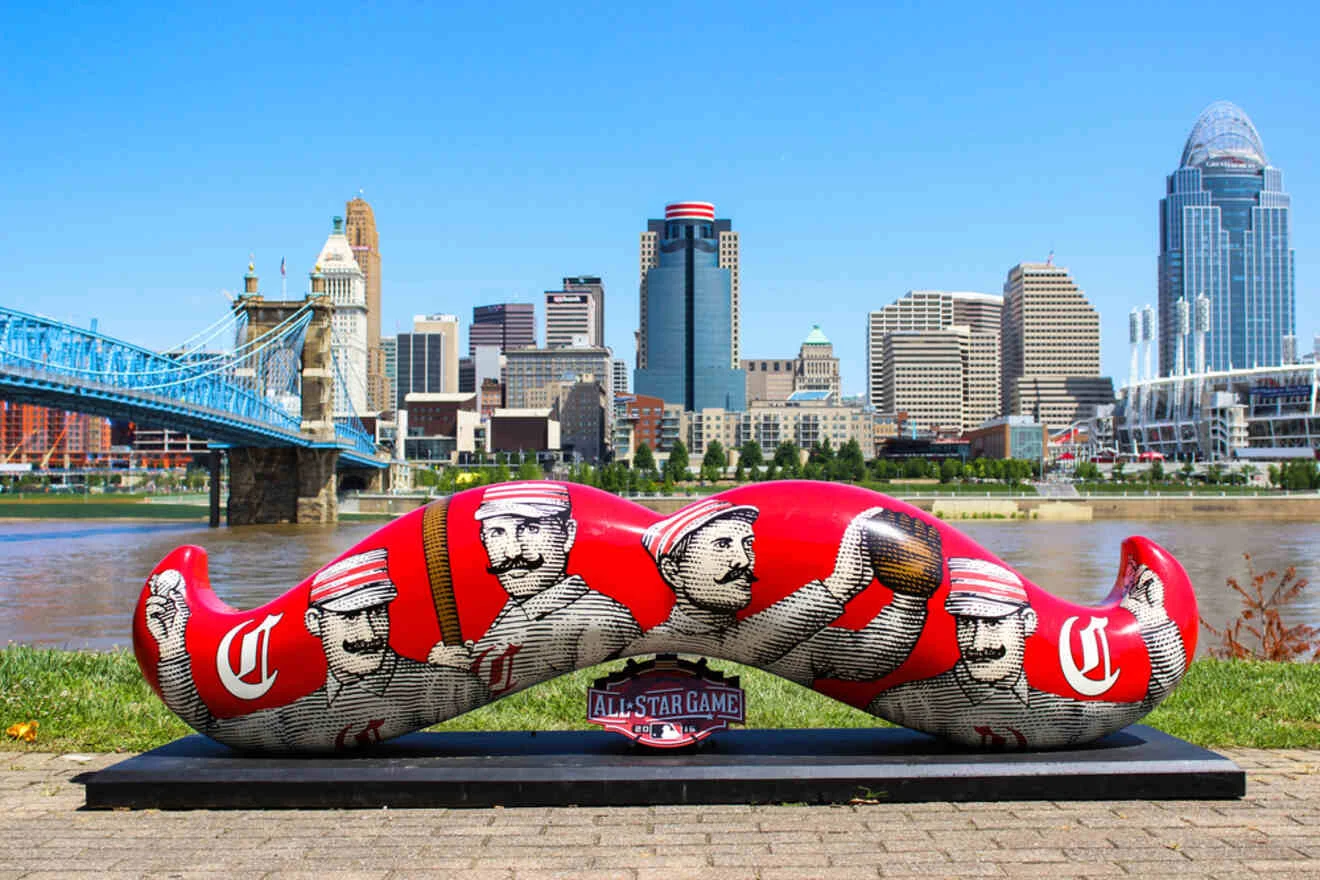 Large red statue of a mustache decorated with vintage baseball player images stands in a park, with a city skyline and river in the background.