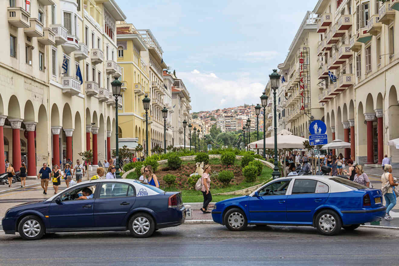 People walk along a bustling street with two blue cars parked beside a garden area, surrounded by historic buildings with arches and balconies.