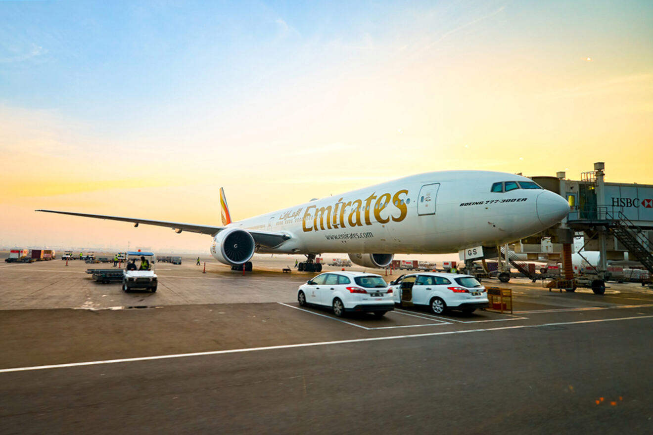 A parked Emirates airplane at an airport gate, with vehicles nearby and the sky in the background.