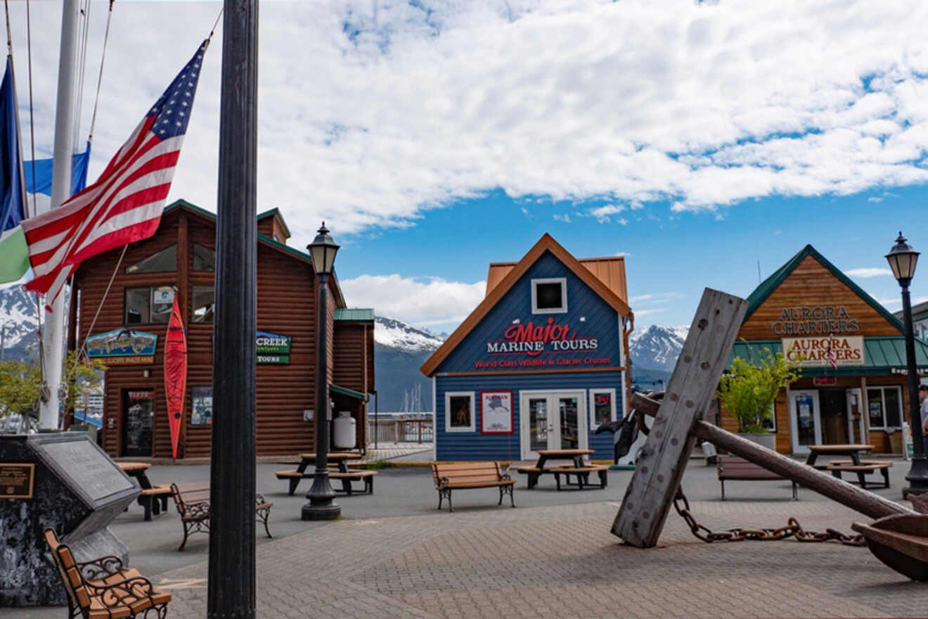 A harbor scene with shops and tours, including Major Marine Tours and Aurora Charters. An anchor sculpture and American flag are in the foreground, with mountains visible in the background.