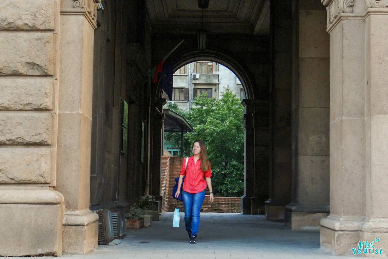 The writer of the post in a red shirt walks under a stone archway holding a shopping bag. There are buildings and greenery in the background.