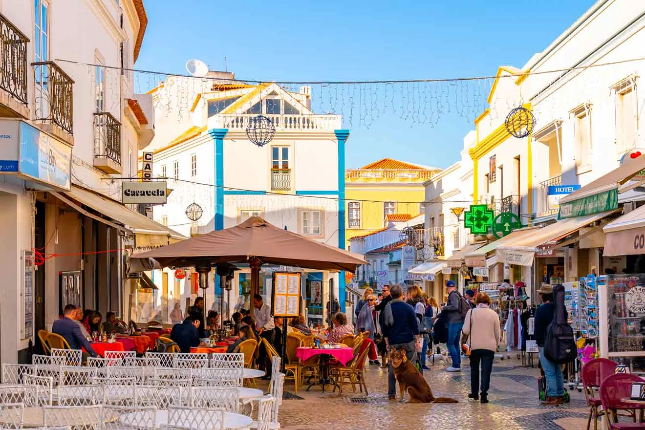 A lively European street scene with people dining at an outdoor café. Shops line the street, and a blue sky is overhead.