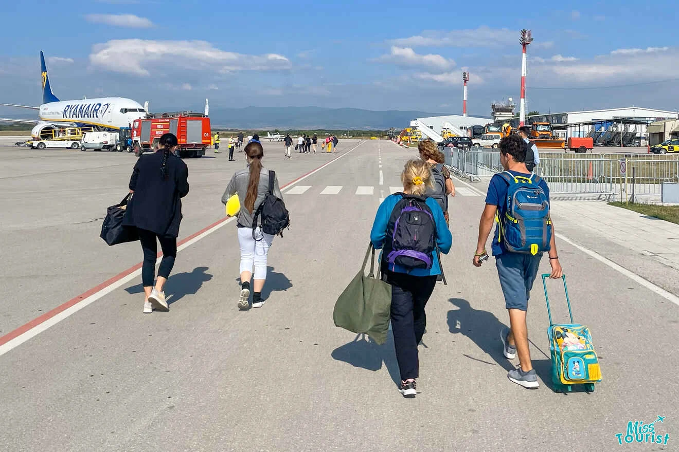 Four people with luggage walk on an airport tarmac towards a Ryanair airplane under a clear sky.