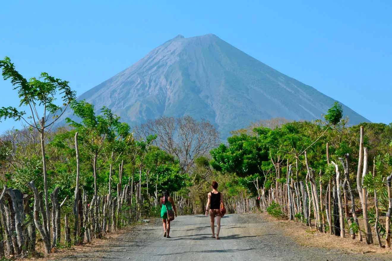 Two people walk down a dirt road surrounded by trees, with a large volcano in the background under a clear blue sky.