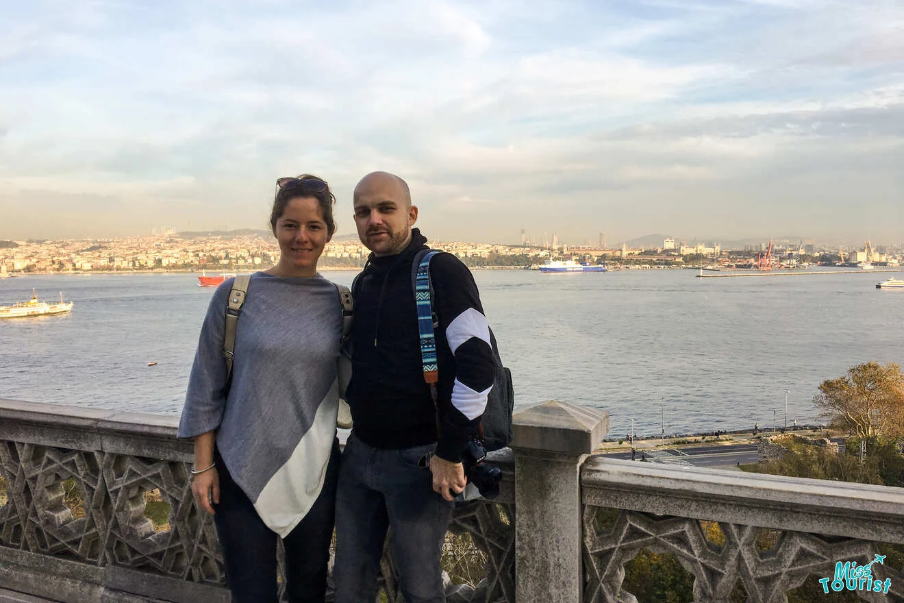 The writer of the post with her husband pose together on a balcony overlooking a body of water with ships. The city skyline is visible in the background.