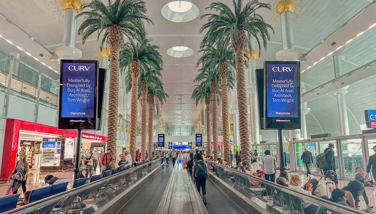 Airport terminal interior with tall palm trees, a moving walkway, and large advertisements. Passengers are walking and using the walkway.