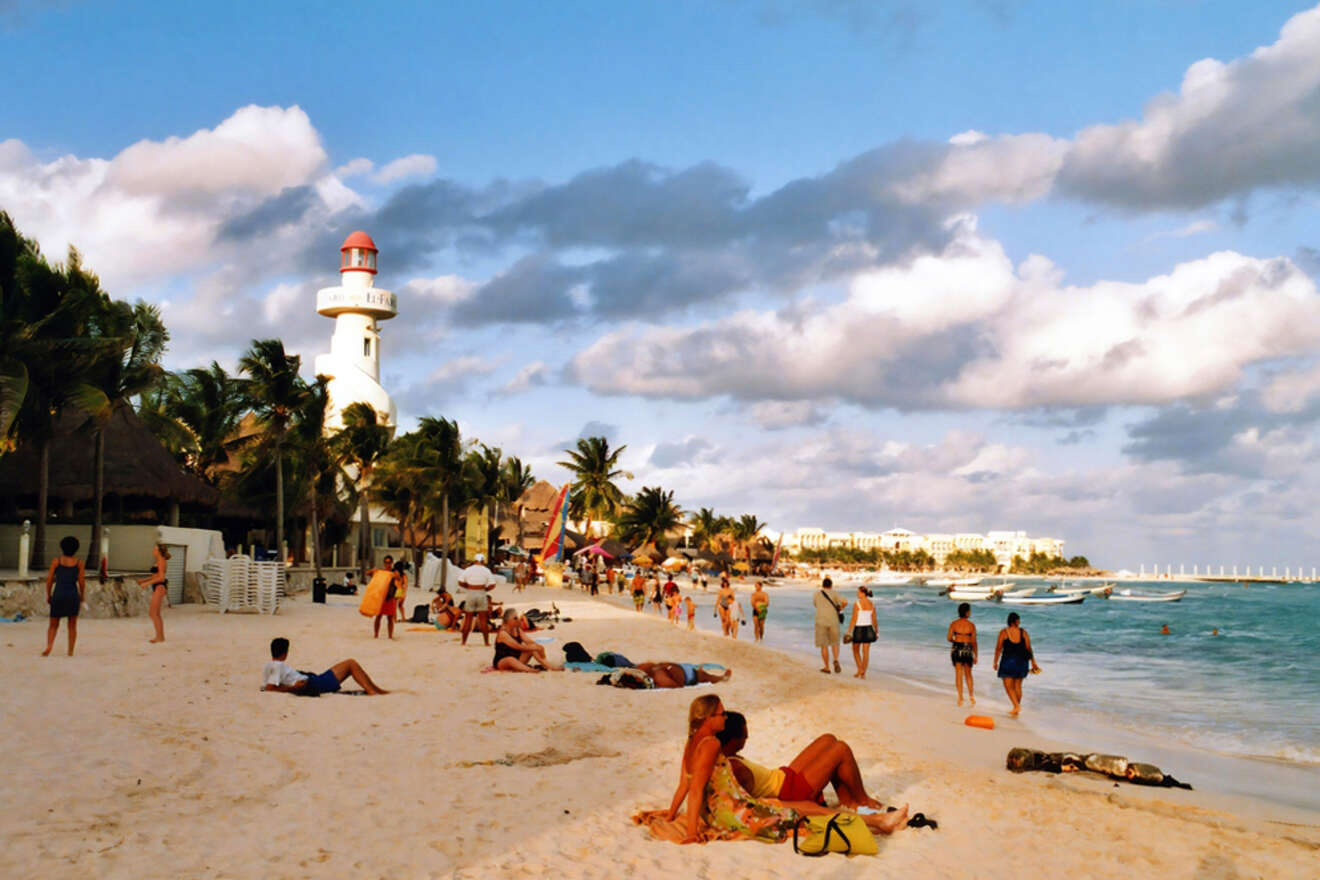 People relax on a sandy beach with a lighthouse, palm trees, and several buildings in the background under a partly cloudy sky.