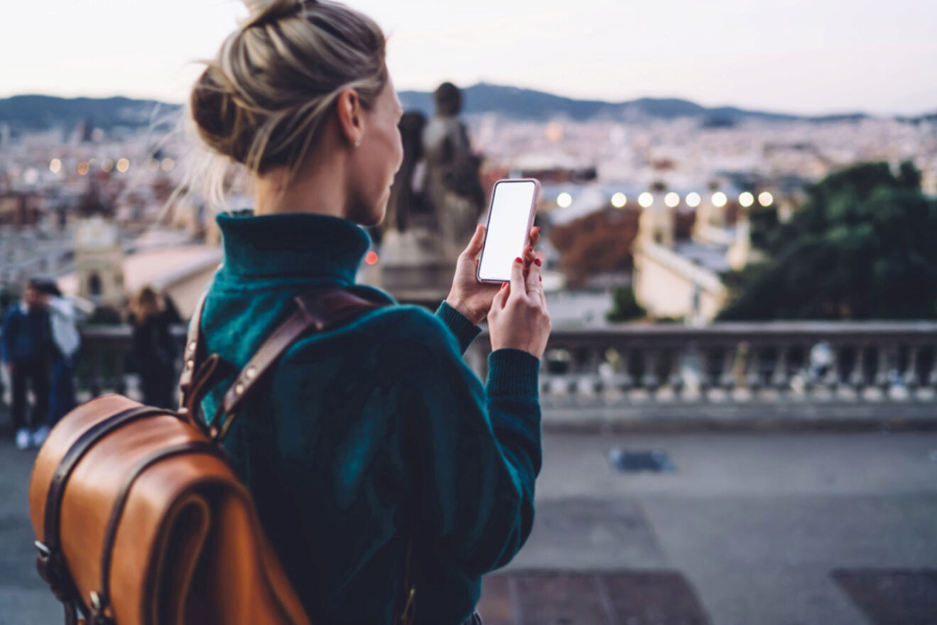 A person with a backpack looks at a smartphone while standing on a terrace overlooking a cityscape.