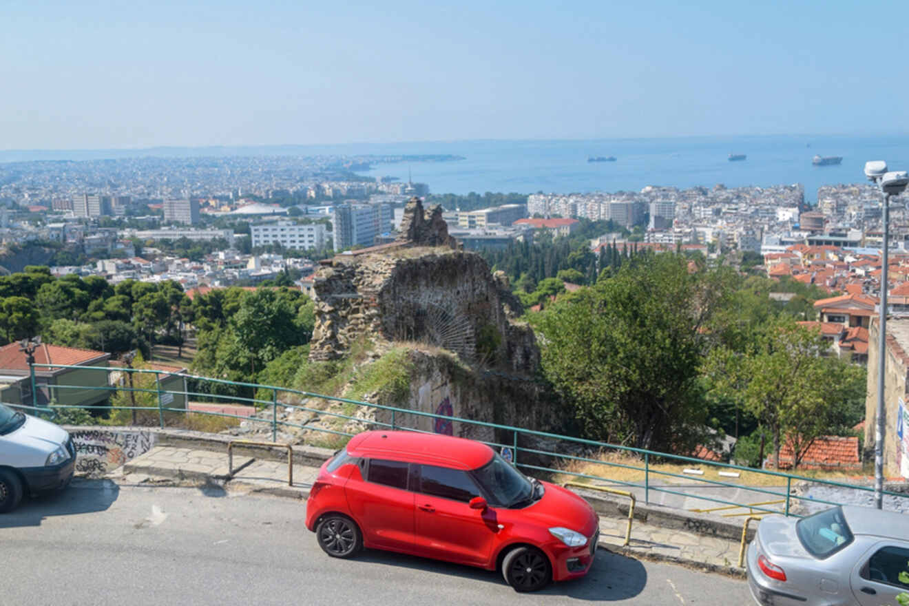 View of a cityscape with a red car in the foreground, ancient ruins, and a coastline with ships in the background under a clear blue sky.