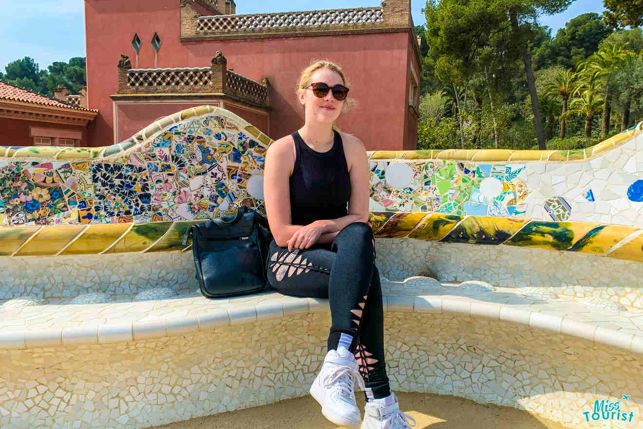 The writer of the post wearing sunglasses sits on a colorful mosaic bench in a sunny outdoor setting with vibrant trees and buildings in the background.