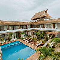 Courtyard view of a tropical resort with a pool, surrounded by lounge chairs, palm trees, and a thatched-roof building.