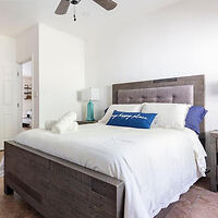 A bedroom with a wooden bed frame, white bedding, and a decorative pillow. A table lamp is on the nightstand, and a ceiling fan hangs above.